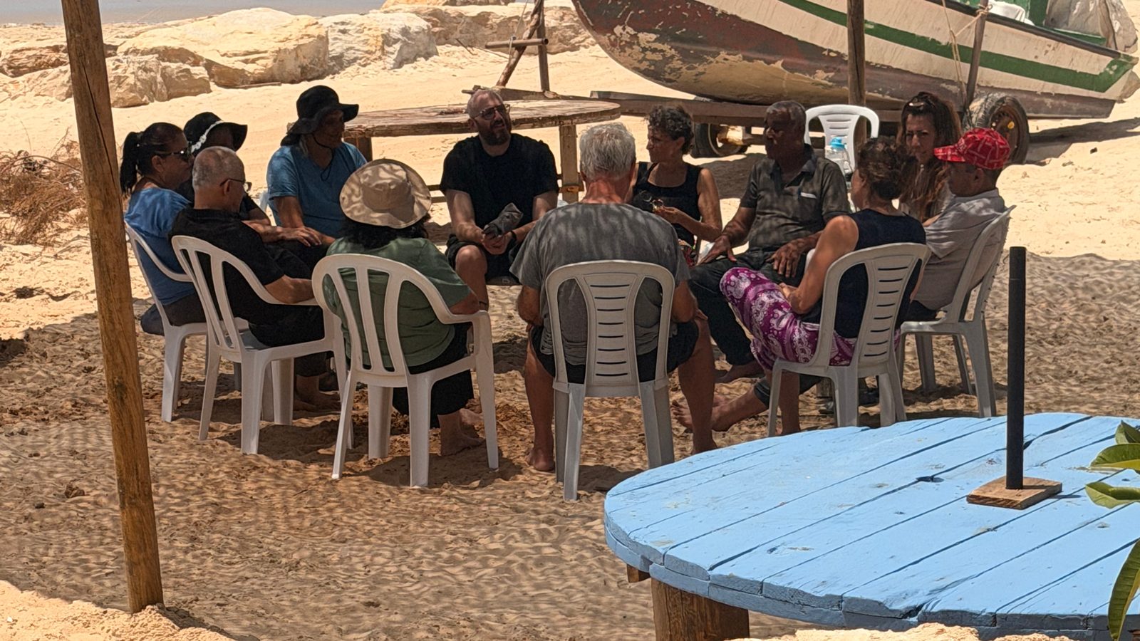 קבוצה יושבת במעגל על החול בצל, ליד סירת דייגים ומול הים