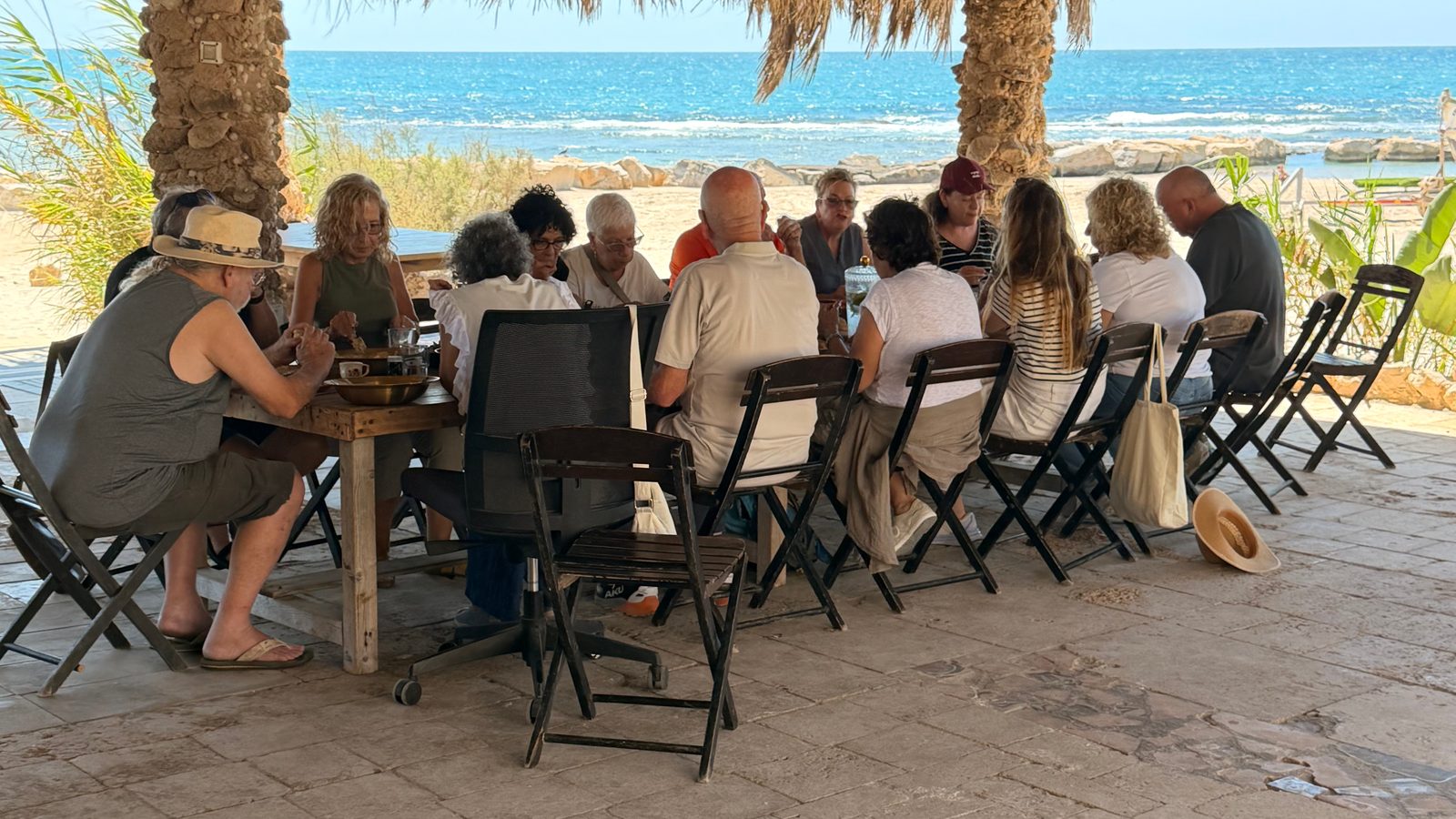 קבוצה יושבת סביב שולחן עבודה ארוך תחת הפרגולה של לב ים, והים מאחוריה